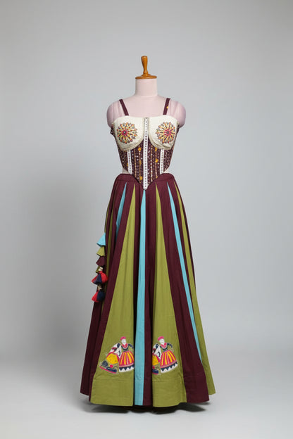 A colorful bohemian-style maxi dress displayed on a mannequin in a clothing store. The dress features a white embroidered bustier top with red and yellow floral patterns, a beaded waist, and a long skirt with vertical stripes of olive green, dark purple, and light blue, decorated with embroidered figures and tassels.