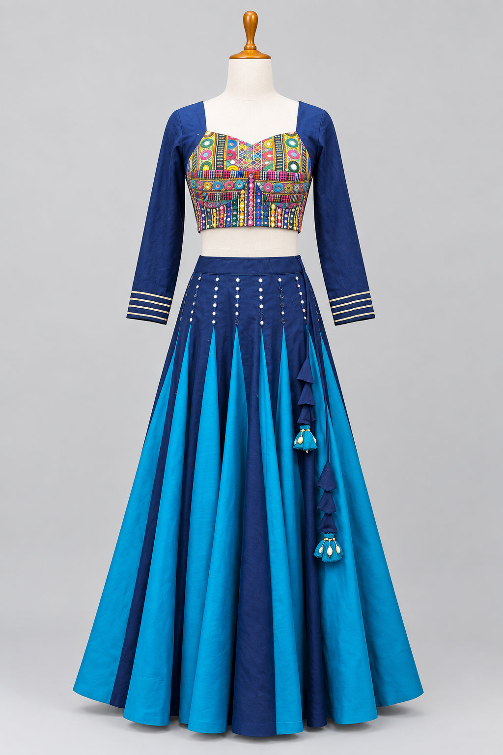 A traditional Indian ethnic wear set displayed on a mannequin in a clothing store. The set features a navy blue long-sleeve kurta with colorful geometric embroidery on the chest and striped cuffs, paired with a matching navy blue and bright blue pleated skirt with tassel details.