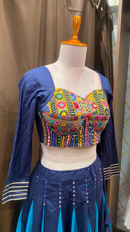 A blue traditional-style outfit displayed on a mannequin, featuring a heavily embroidered multicolor bustier-style top with a white underlayer and a blue skirt with turquoise panels and striped sleeve cuffs.